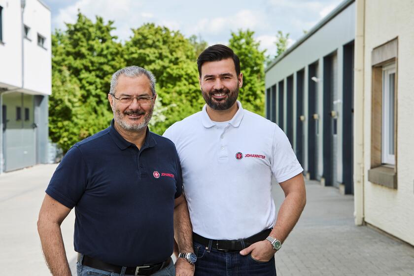 Ein älterer Mann mit blauem Johanniter-Polo-Shirt und ein jüngerer Mann im weißen Polo-Shirt auf einem Platz zwischen zwei Gebäuden