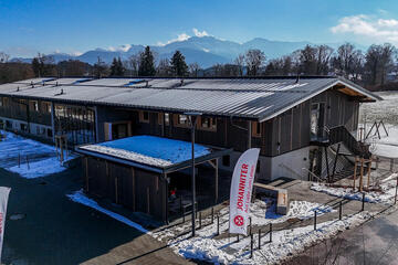 Luftaufnahme: Gebäudekomplex am Feldrand, verschneite Landschaft und Alpenblick.