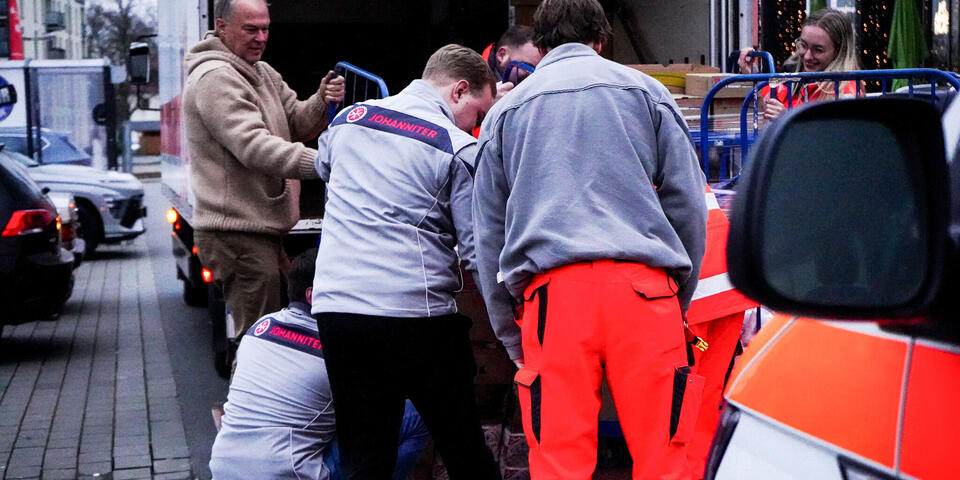 Männer und Frauen, teilweise in Johanniter-Kleidung, schieben einen gefüllten Rollbehälter auf die Rampe eines LKW