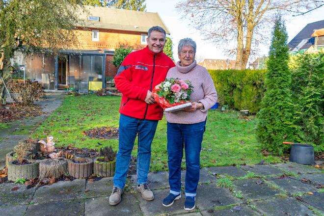 Ein Mitarbeiter der Johanniter und eine Seniorin stehen in einem Garten und lächeln in die Kamera.