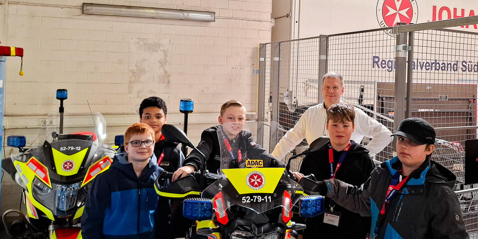Ein Junge auf einem Johanniter-Motorrad. Daneben stehen weitere Jungen und ein Mann