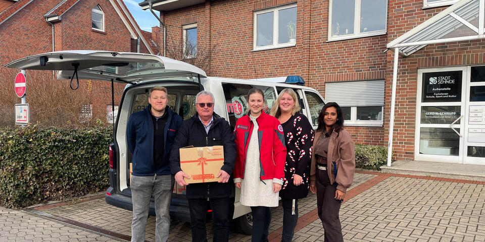 Ein Mann mit einem Paket. Neben ihm stehen zwei Frauen und eine Frau und ein Mann mit einer Johanniter-Jacke. Im Hintergrund ein Transporter mit geöffneter Heckklappe