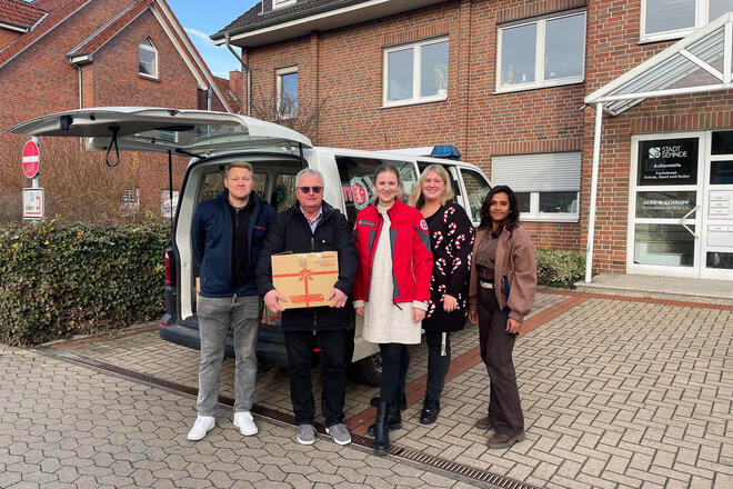 Ein Mann mit einem Paket. Neben ihm stehen zwei Frauen und eine Frau und ein Mann mit einer Johanniter-Jacke. Im Hintergrund ein Transporter mit geöffneter Heckklappe