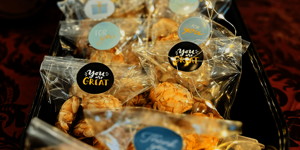 Mehrere durchsichtige Tütchen, gefüllt mit Plätzchen, liegen auf einem Tablett – jedes Tütchen ist mit einem bunten Aufkleber wie „You are GREAT“ oder „For you“ versehen.