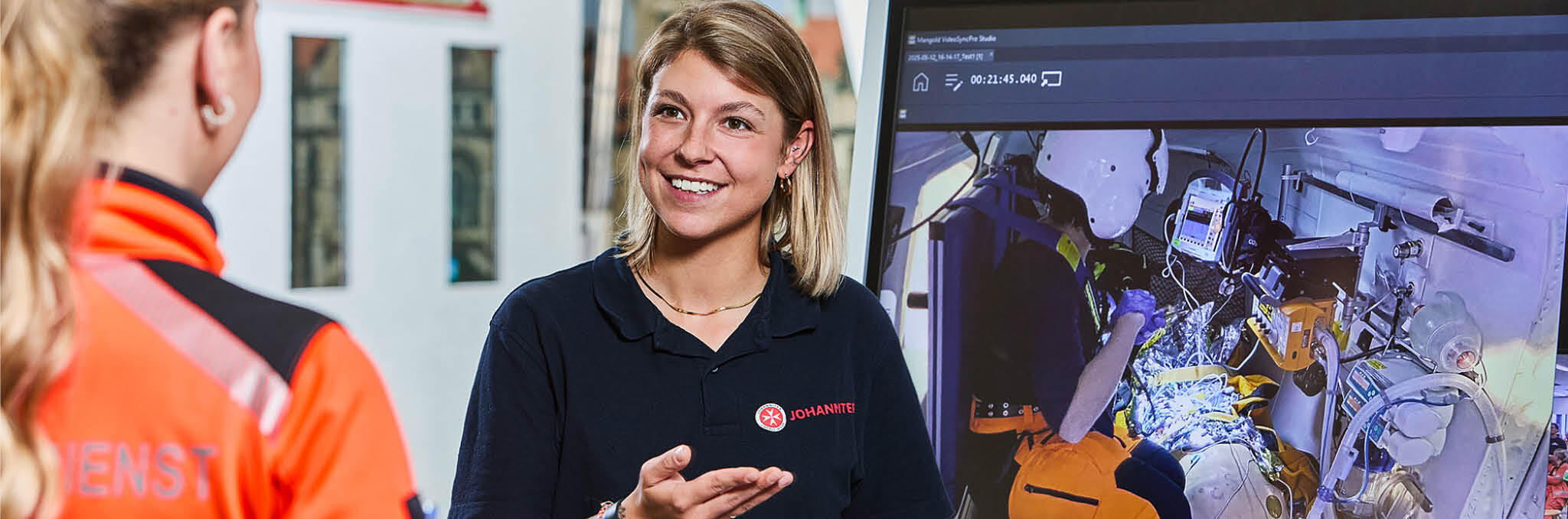 Eine Dozentin steht vor einem großen Monitor in der Rettungsschule und erklärt einer Rettungssanitäterin etwas.