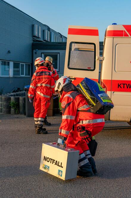 Drei Johanniter in Einsatzkleidung stehen vor einem Krankenwagen. Eine Person hebt einen Notfallkoffer vom Boden auf.