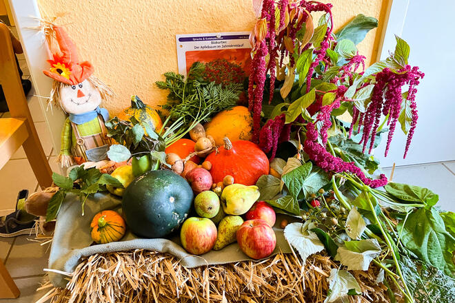 Ein mit Gemüse, Obst, Stroh und Blumen dekorierter Tisch. Links ist eine Vogelscheuchenpuppe