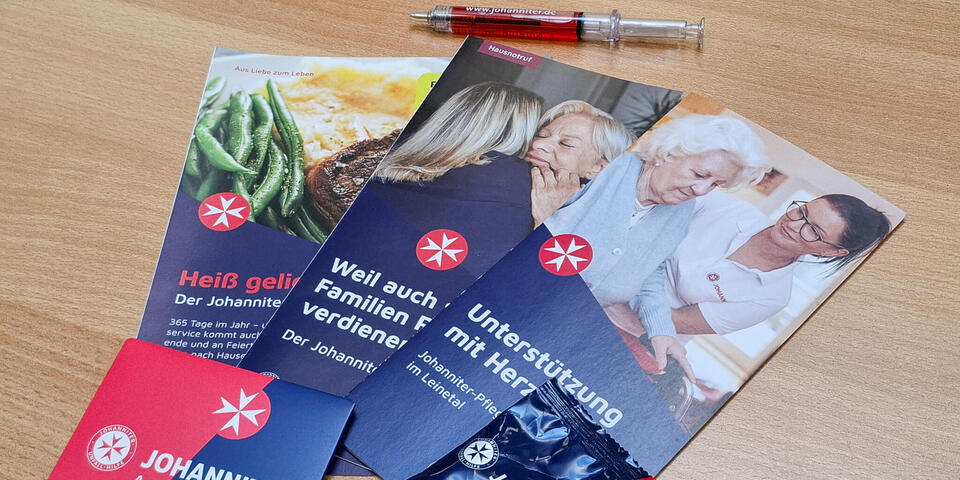 Drei Johanniterflyer zu Menüservice, Hausnotruf und Pflege, ein Kugelschreiber in Spritzenform, Gummibärchen und ein Pflastermäppchen auf einem Tisch.