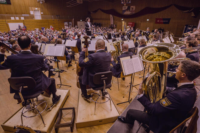 Musiker in Uniformen geben Konzert vor voll besetzter Tribüne in einer Mehrzweckhalle.