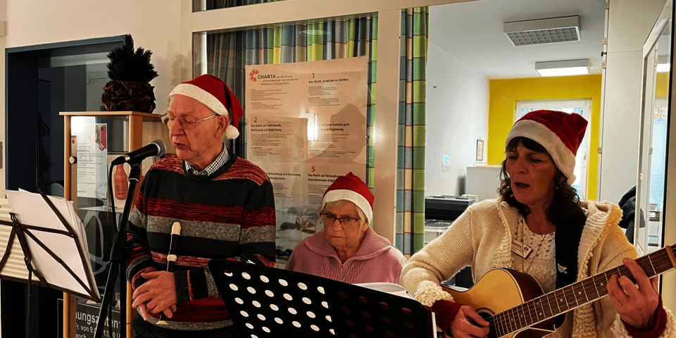 drei Personen mit Weihnachtsmützen stehen singen und mit Instrumenten hinter Notenständern