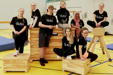 Acht junge Ferienbetreuerinnen und -betreuer posieren fröhlich in einer Turnhalle. Sie sind in sportlicher Kleidung und tragen alle einheitliche "Lilalu"-T-Shirts