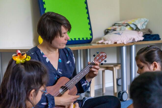 Musikkurs in der Kinderbetreuung Ostseestraße Eine Frau spielt eine Ukulele, im Vordergrunds sitzen Kinder.