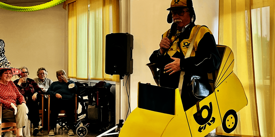Ein Mann in einem Kostüm, das wie ein kleines gelbes Postauto aussieht, hält ein Mikrofon in der Hand.