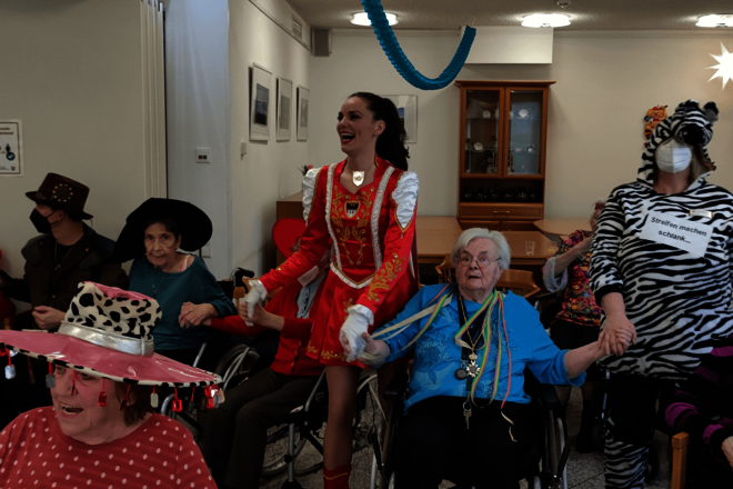 Ein rotgekleidetes Tanzmariechen hält die Hände der neben ihr sitzenden Senioren.
