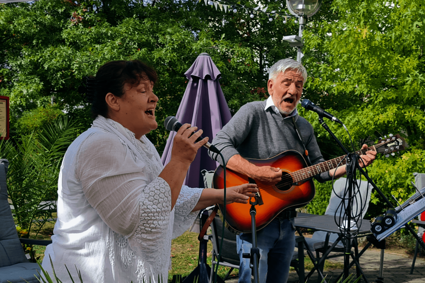 Eine Frau in weißer Bluse hält auf einer Terrasse im Garten ein Mikrofon in der Hand und singt. Daneben spielt ein Mann in einem grauen Pullover Gitarre und singt ebenfalls.