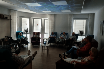 In bequemen Ruhesesseln hört eine Gruppe von Senioren eine vorlesenden Frau zu.