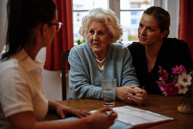 drei Frauen sprechen am Tisch miteinander: Eine ältere Dame, eine Angehörige und eine Pflegekraft in weißer Berufskleidung