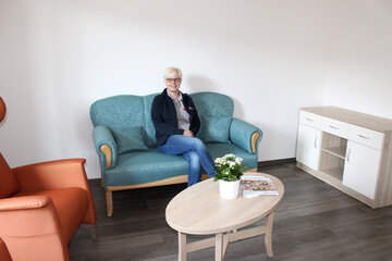 Carmen Birkholtz sieht im gemütlichen Wohnbereich der WG auf dem Sofa und lächelt in die Kamera. 