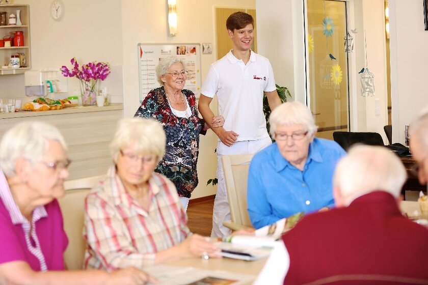Ein Pfleger führt eine lachende Seniorin an einen Tisch, an dem schon andere Damen und Herren sitzen.
