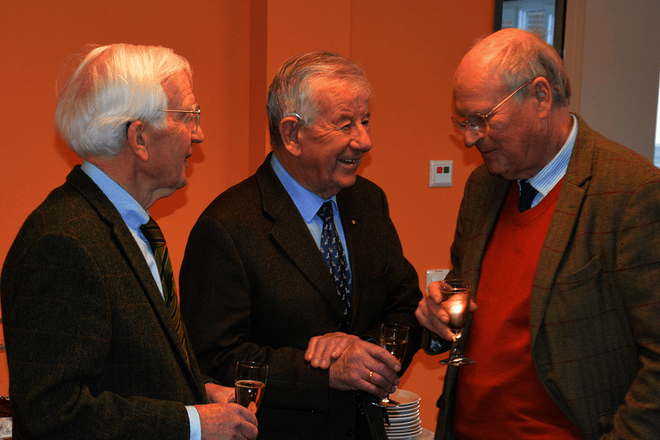 Drei ältere Herren unterhalten sich. Sie halten Seltgläser in den Händen. Im Hintergrund ein kaltes Büffet.