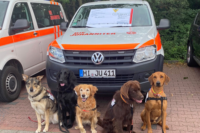 4 Hunde sitzen vor einem Einsatzfahrzeug auf dem ein symbolischer Scheck liegt