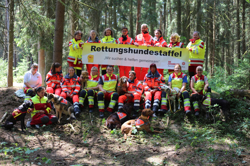 Einsatzkräfte mit Hunden halten ein Plakat mit der Aufschrift "Rettungshundestaffel - Wir suchen und helfen gemeinsam" hoch