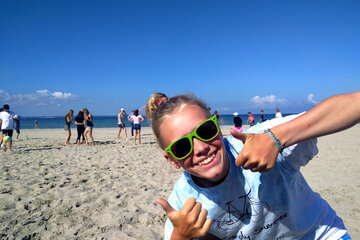 Ein Mädchen mit Sonnenbrille am Strand guckt in die Kamera und zeigt beide Daumen hoch.