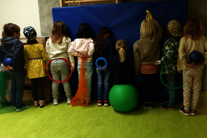 Mehrere Kinder stehen mit dem Rücken zum Betrachter vor einer blauen Sportmatte. In den Händen halten sie verschiedene Reifen.