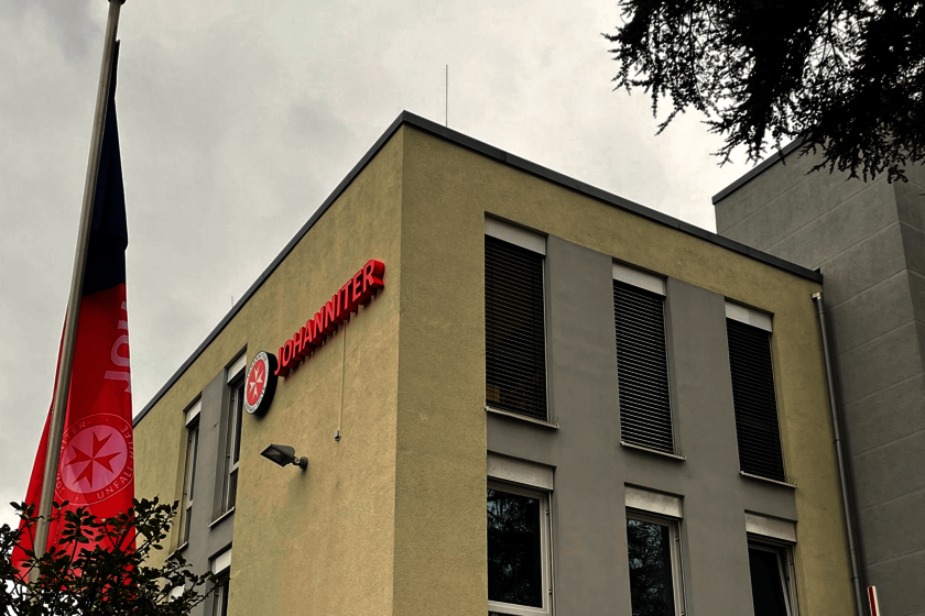 mehrstöckiges Gebäude mit Johanniter-Logo und rot-blauen Fahnen im Vordergrund