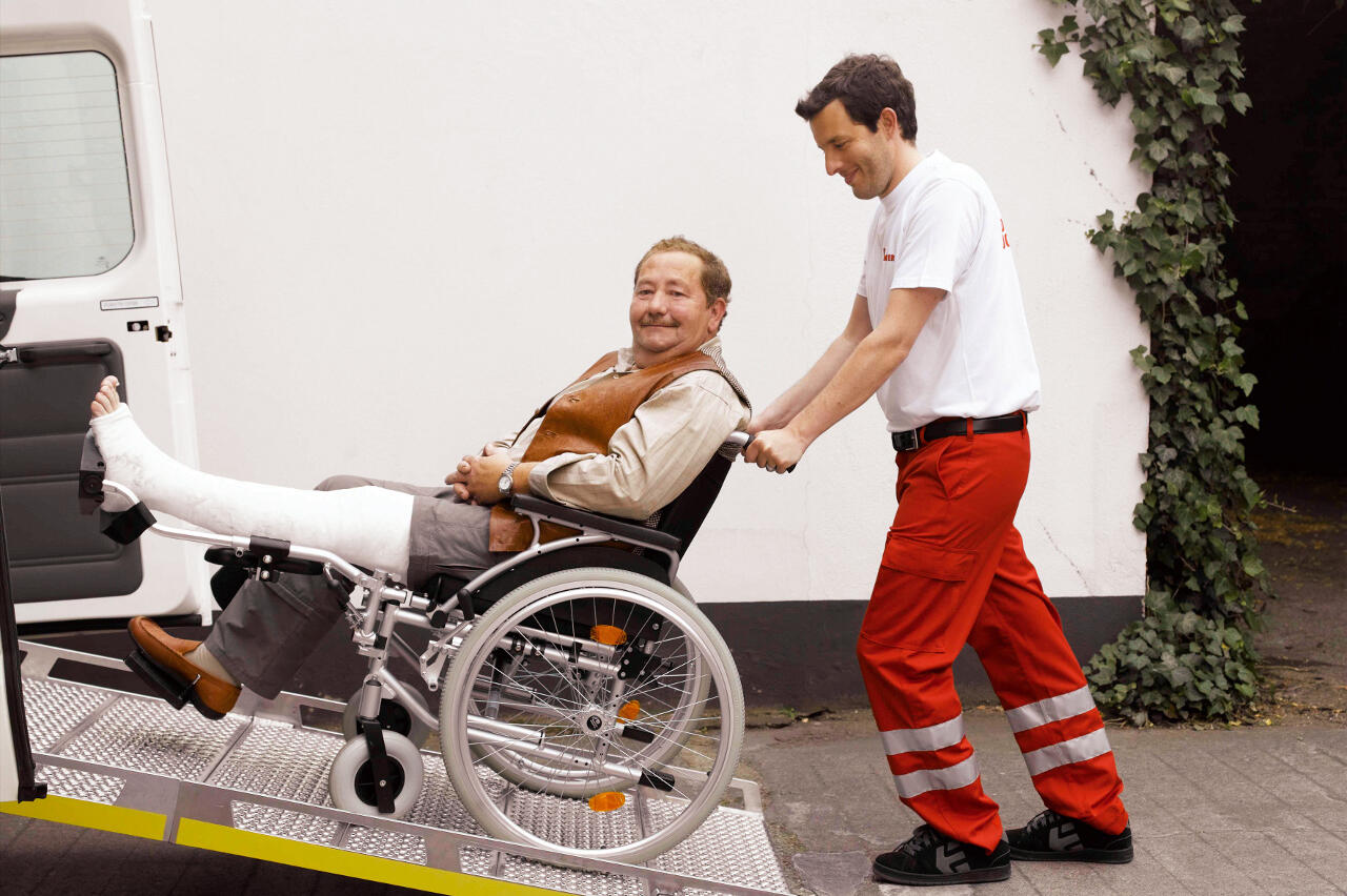 Ein Mitarbeiter des Johanniter-Fahrdienstes schiebt einen Mann mit Gips am Fuß im Rollstuhl in ein Auto.
