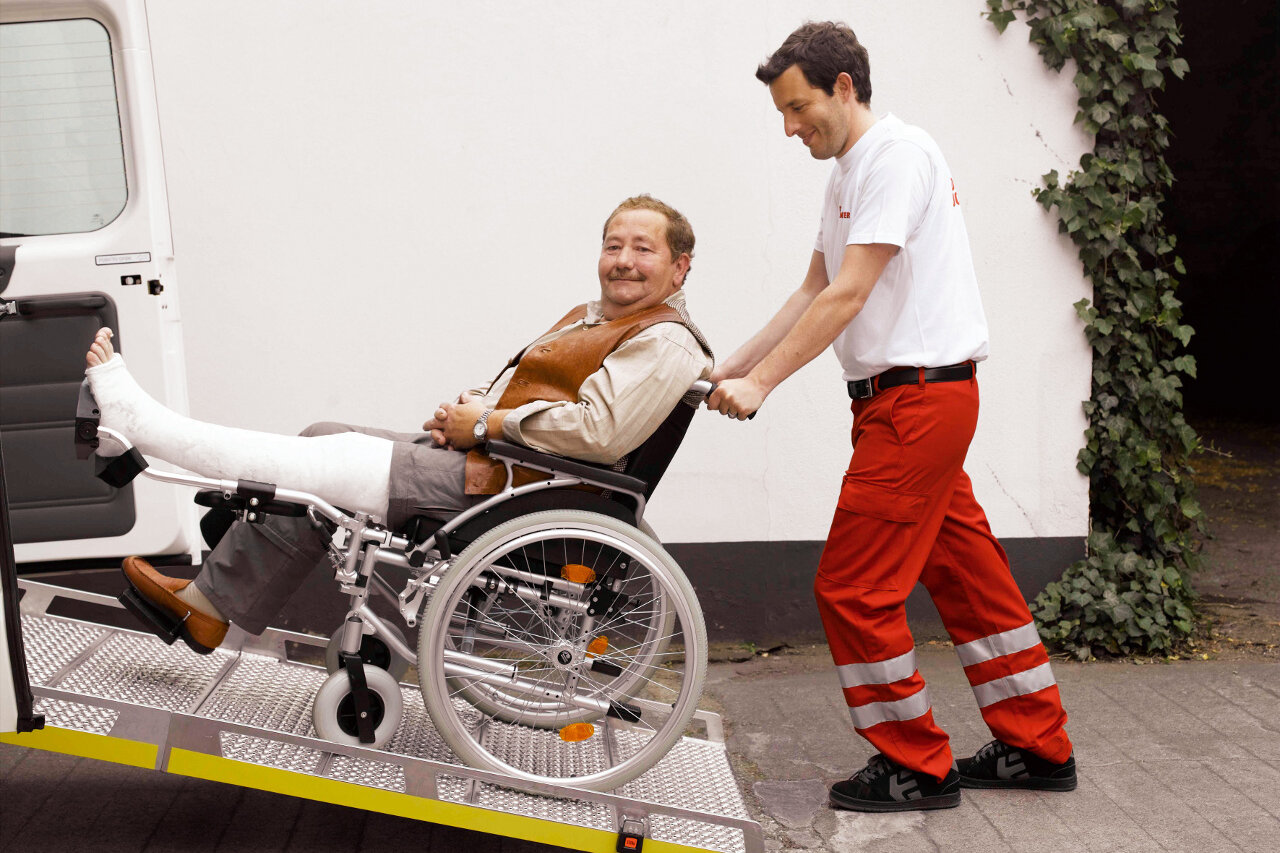 Ein Mitarbeiter des Johanniter-Fahrdienstes schiebt einen Mann mit Gips am Fuß im Rollstuhl in ein Auto.