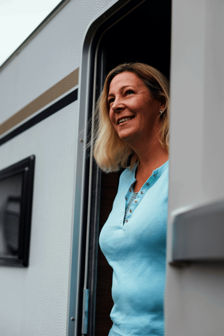 eine blonde Frau in türkisem T-Shirt lächelt aus der Tür eines Wohnwagens.