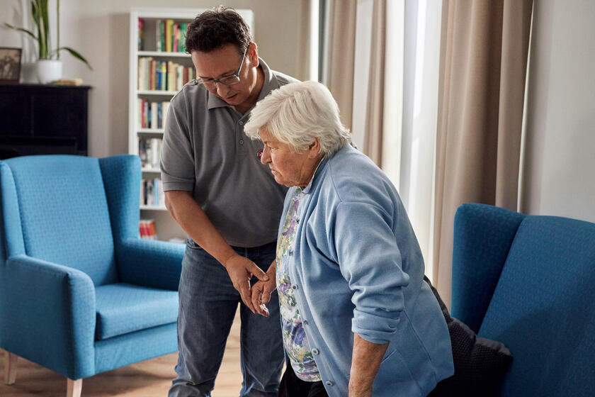 Ein Mitarbeiter aus der Pflege hilft einer Seniorin beim Aufstehen.