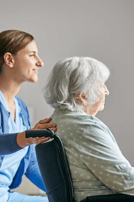Eine Pflegerin legt einer älteren Frau im Rollstuhl die Hand auf den Rücken.
