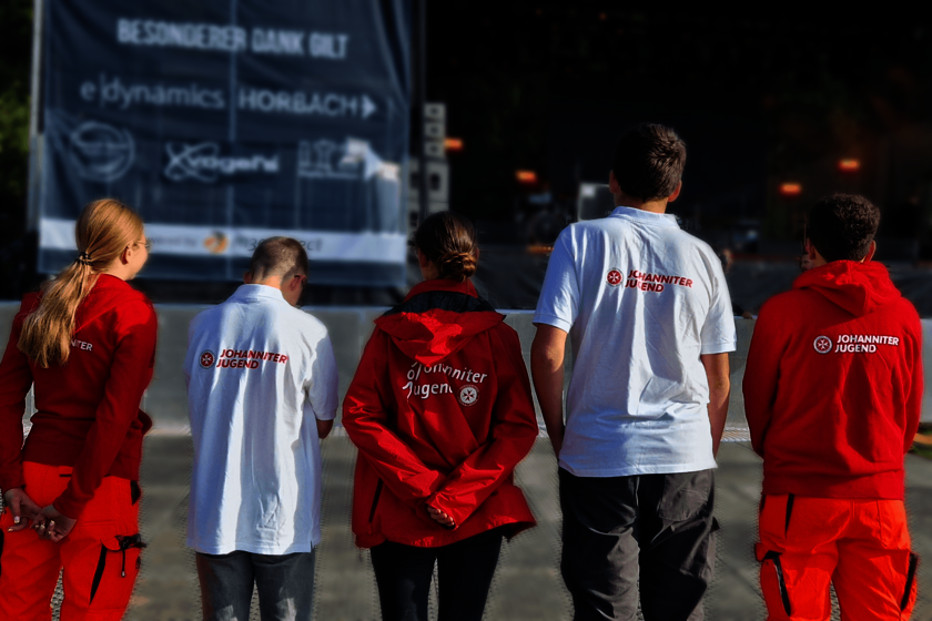 Fünf junge Menschen stehen nebeneinander auf einem großen Platz vor einer Bühne. Sie sind von hinten zu sehen und tragen weiße Shirts oder rote Swetshirts und Jacken. Auf allen Rücken ist das Logo der Johanniter-Jugend zu sehen.