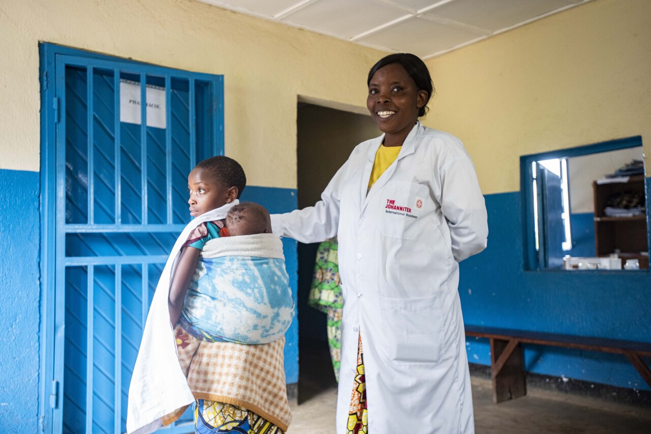 Krankenschwester in Gesundheitsstation in Nord-Kivu berührt lächelnd ein junges Kind auf dem Rücken der Mutter