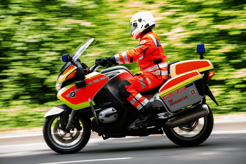Eine Person in leuchtendroter Einsatzkleidung sitzt auf einem Einsatzmotorrad vor einem Hintergrund aus Büschen. Der Foto zeigt durch Bewegungsunschärfe, dass das Fahrzeug in Bewegung ist.