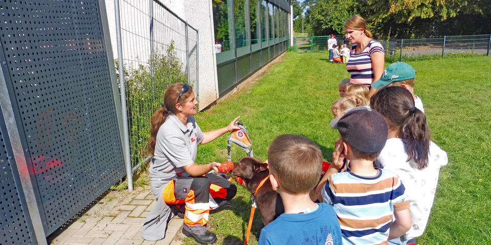 Eine Johanniterin mit Hund zeigt Kindern eine Kenndecke für Rettungshunde