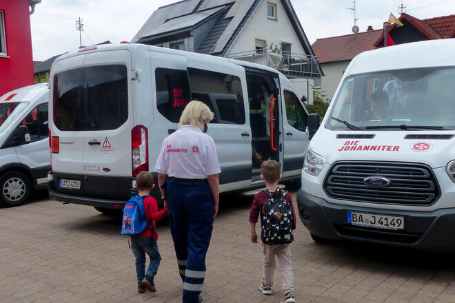 Eine Mitarbeiterin des Johanniter-Linienfahrdienstes begleitet zwei Kinder zum Bus.