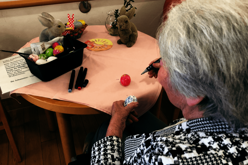 Ältere Dame sitzt an einem österlich geschmückten Tisch und bemalt ein Osterei.