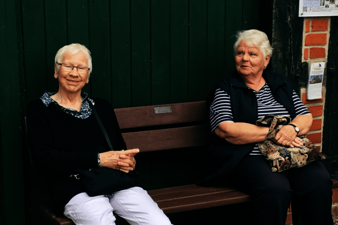 Zwei Frauen sitzen vor einer grüngestrichenen Holzwand auf einer Bank.