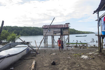Ein Boot, ein Haus und zwei Kinder auf matschigem Untergrund, im Hintergrund Wasser