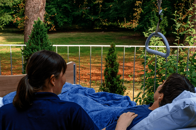 Ein Mensch mit kurzen dunklen Haaren liegt auf einer Terrasse in einem Bett und sieht in den Garten. Neben sitzt eine Frau in einem blauen Poloshirt und berührt seine Schulter.
