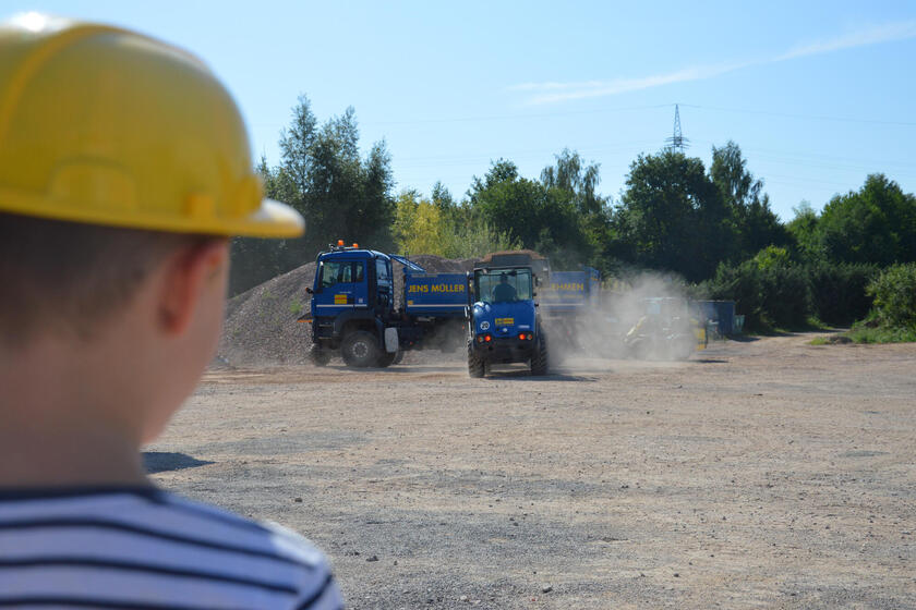 Ein Kind mit einem Bauhelm schaut Einem Bagger zu, der Sand in einen LKW lädt