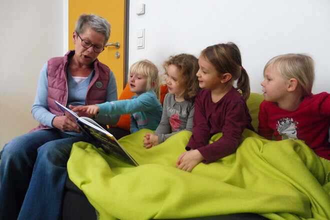 Eine Lesepatin sitzt mit den Kindern auf dem Sofa und liest ihnen vor. Alle Kinder sind unter eine hellgrüne Wolldecke gekuschelt und schauen mit ins das Buch hinein. 