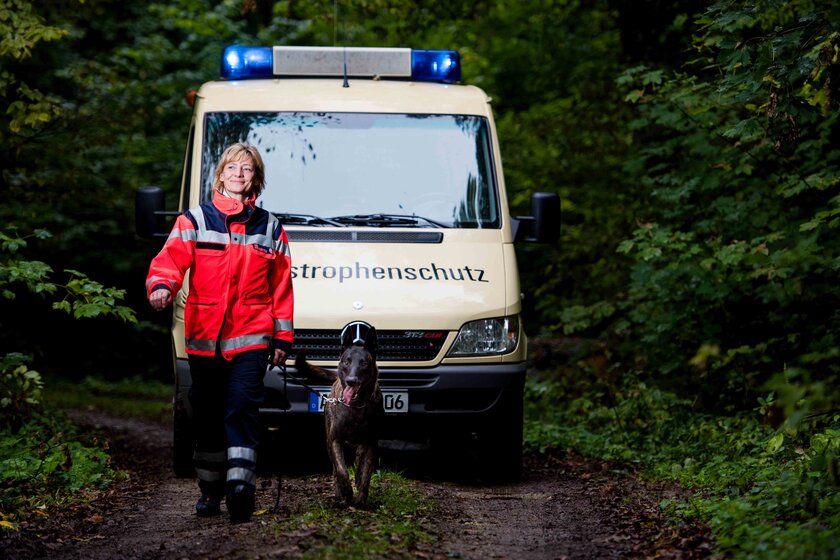 Ein Rettungshundeteam läuft im Wald. Dahinter fährt ein Auto des Katastrophenschutzes.