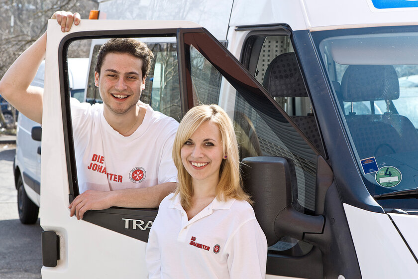Ein junger Mann und eine junge Frau, die ein Freiwilliges Soziales Jahr bei den Johannitern, machen neben einem Dienstauto der Johanniter.