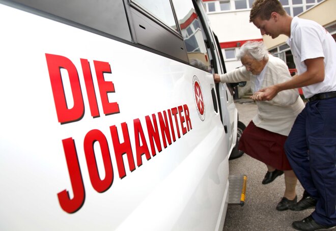 Ein Mitarbeiter des Fahrdiensts der Johanniter-Unfall-Hilfe hilft einer älteren Frau beim Einsteigen in ein Auto.