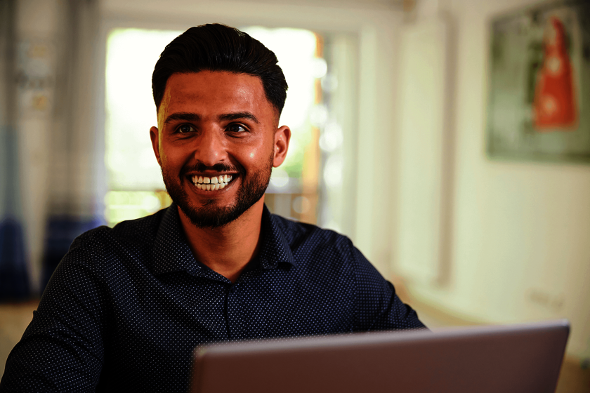 Ein lächelnder junger Mann mit Bart und dunklem Hemd trägt ein dunkelblaues Hemd und sitzt hinter einem Laptop