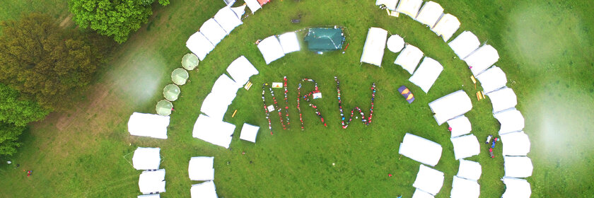 Eine Luftaufnahme von Zelten, die im Kreis angeordnet sind. In der Mitte bilden Menschen die Buchstaben N R W.